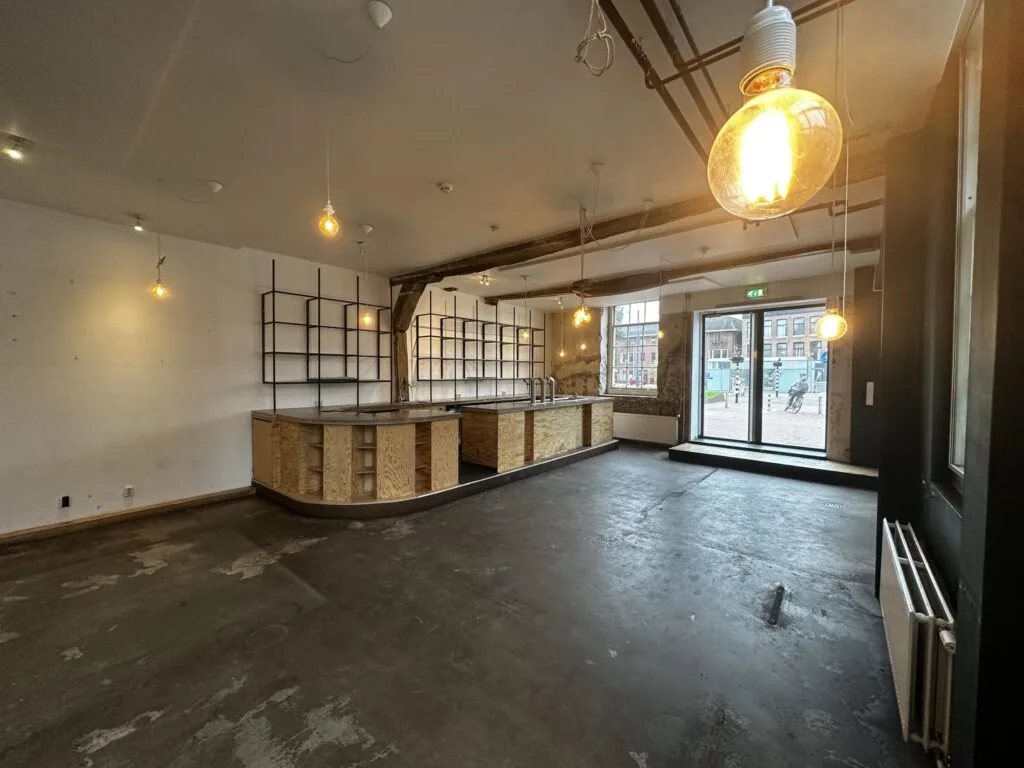 Leegstaand horecapand met industriële uitstraling aan het Sint-Agathaplein, met zicht op de straat door grote ramen.